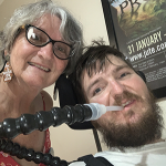 A woman and a bearded man who is wearing a medical breathing tube to clear his throat 